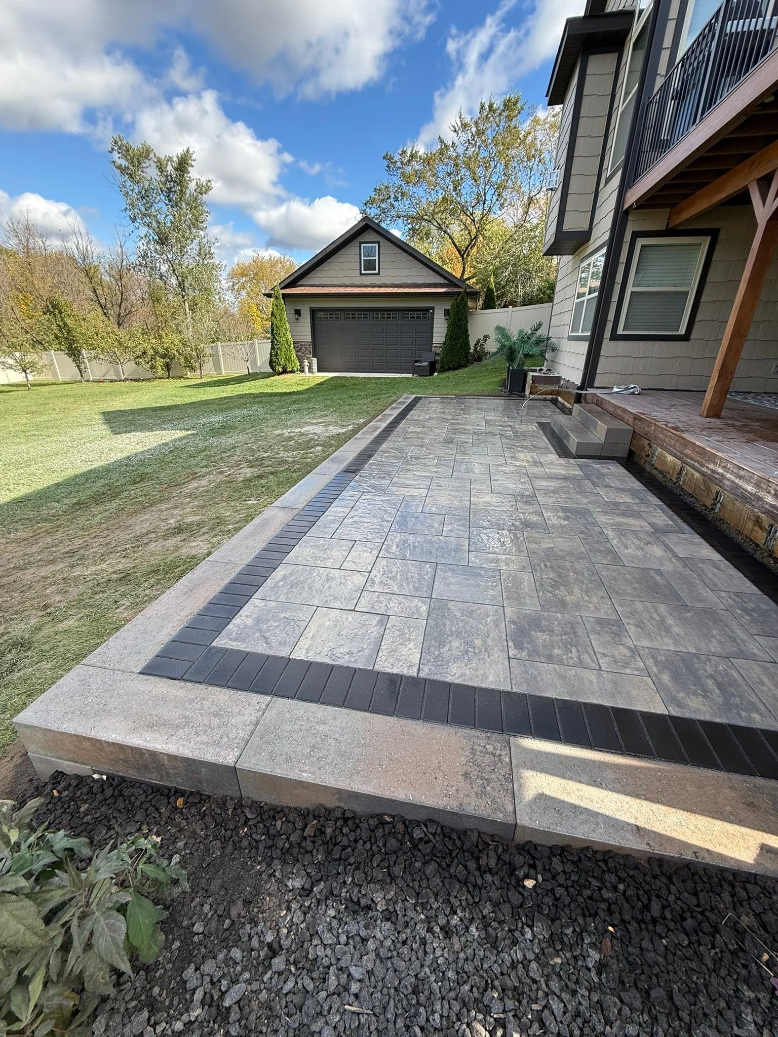Concrete patio beside a brick house and garage, with a grassy yard and trees under a cloudy sky