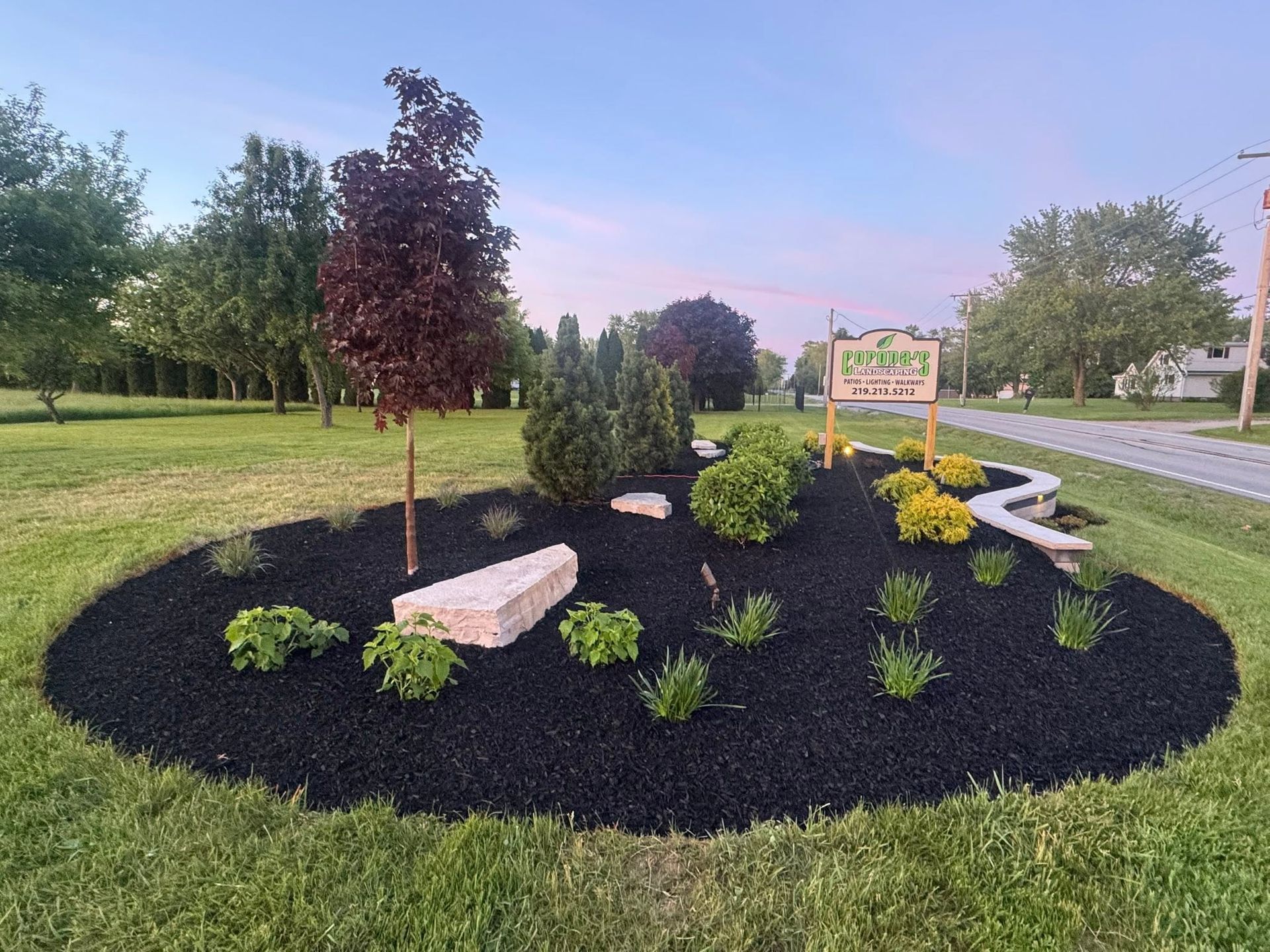 Landscaped garden bed with black mulch, shrubs, a bench, and a sign in a park along a road