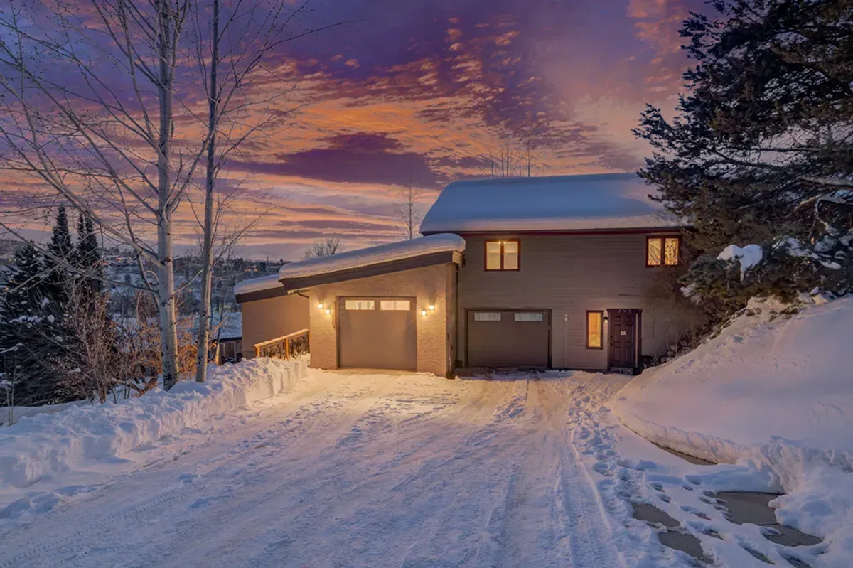 The house is covered in snow and there is a snowy driveway leading to it.