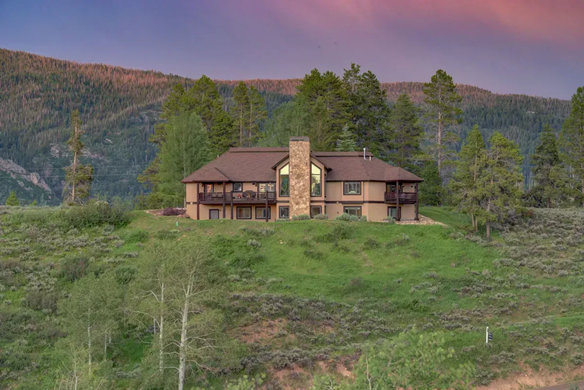 A large house is sitting on top of a grassy hill surrounded by trees.
