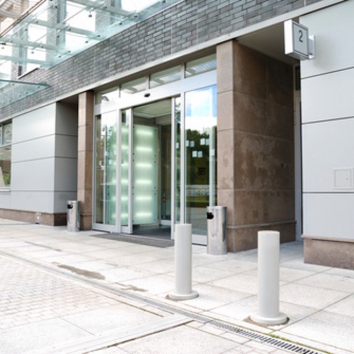 Modern building entrance with glass sliding doors, stone columns, and bollards on a paved walkway