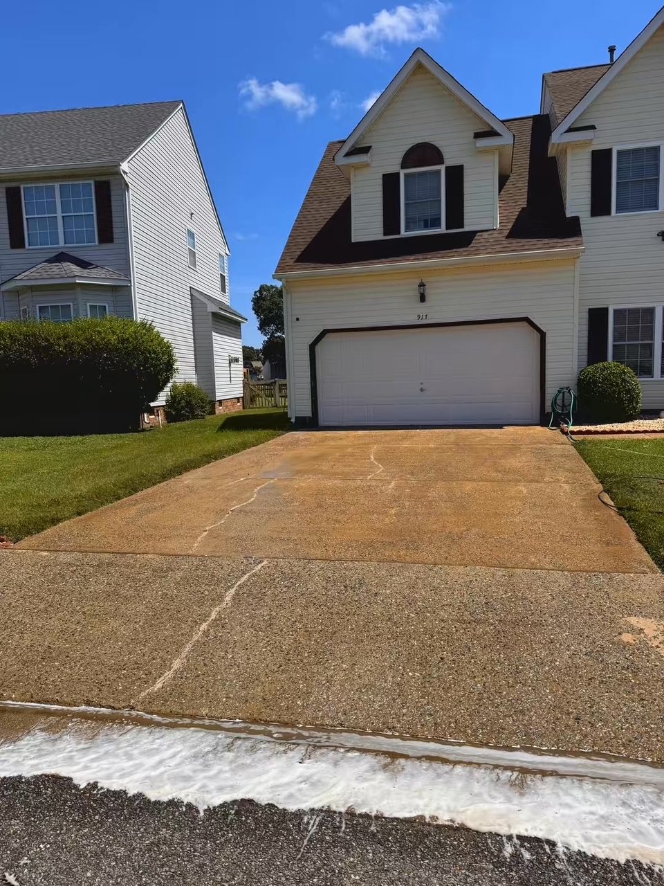 Driveway leading to a two-story house with a white garage door, surrounded by green grass and a blue sky.