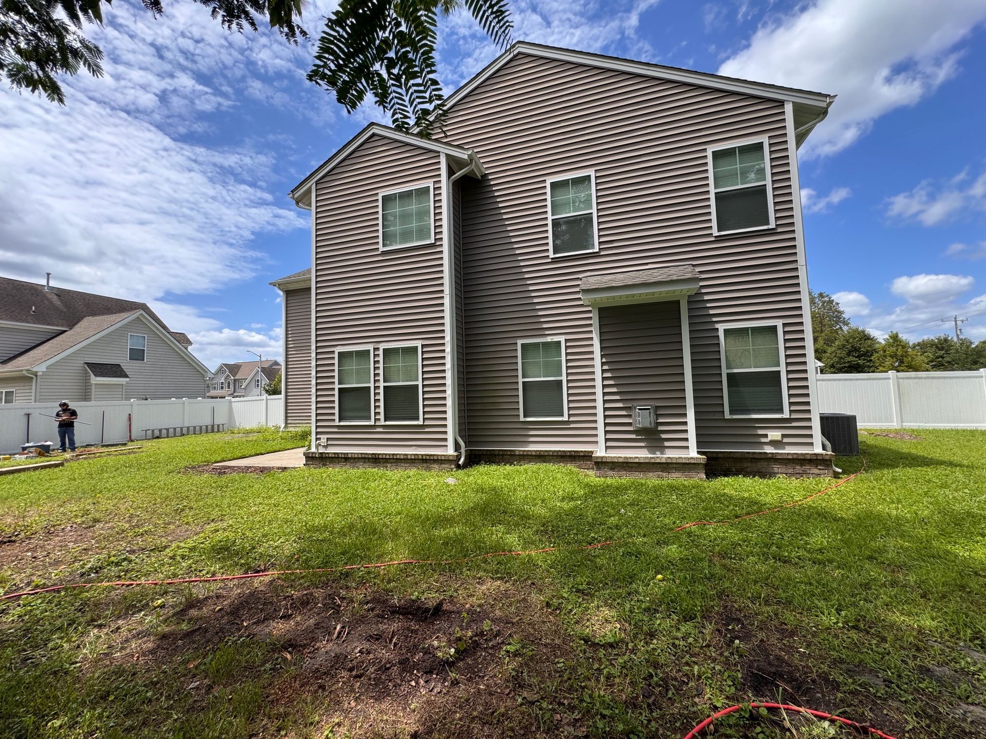 Two-story house with gray siding, several windows, and a grassy yard. A white fence is in the background.
