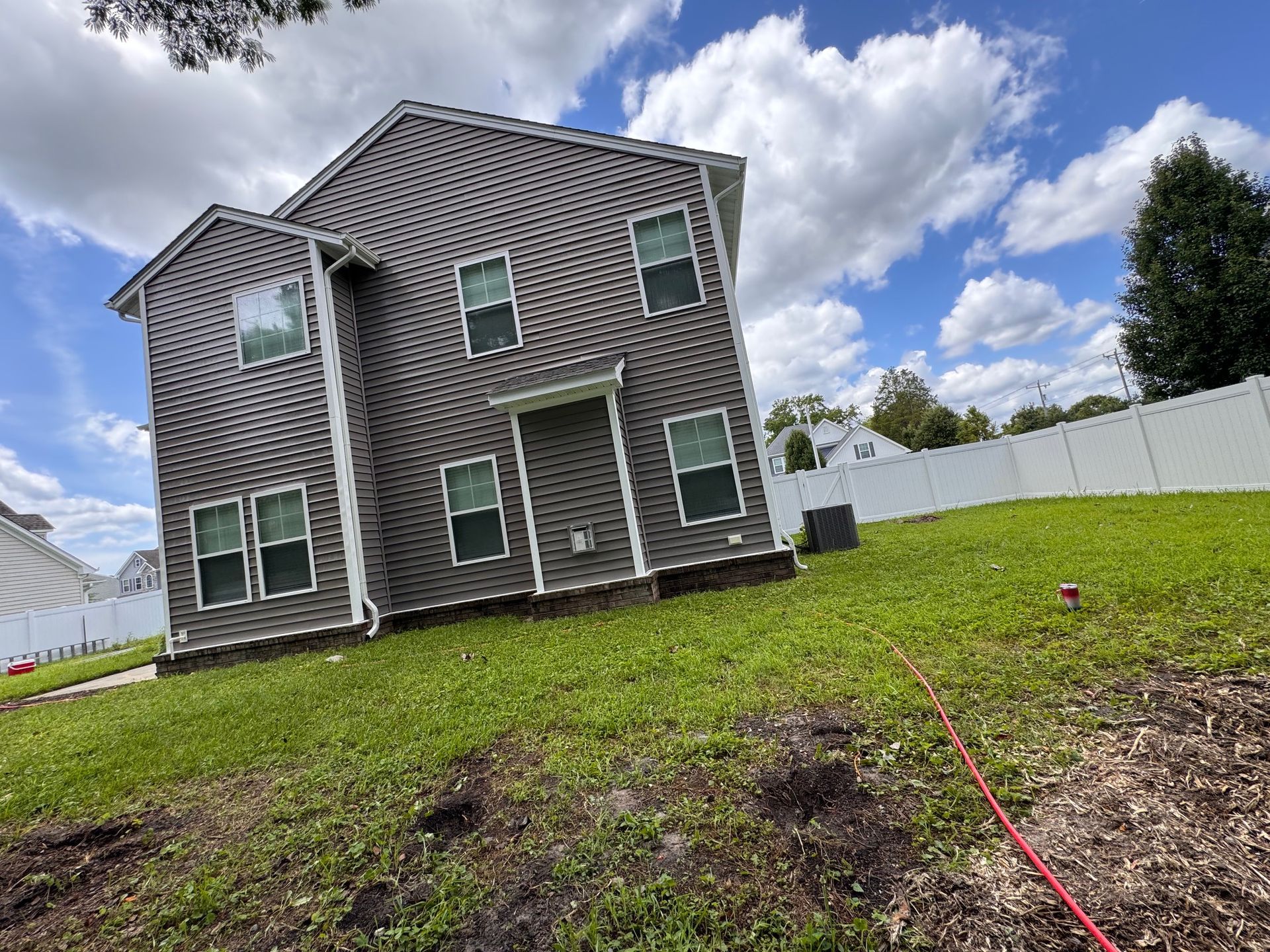 Two-story house with dark siding on a grassy hill, white fence, and partly cloudy sky.