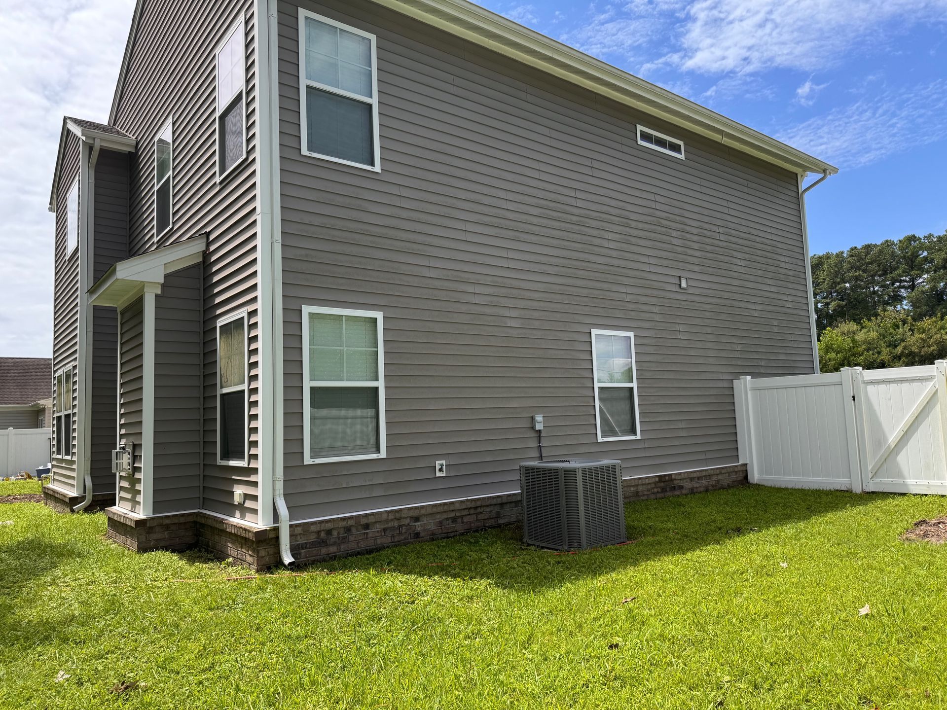 Side of a gray house with white trim, windows, and an air conditioning unit on green grass beside a white fence.