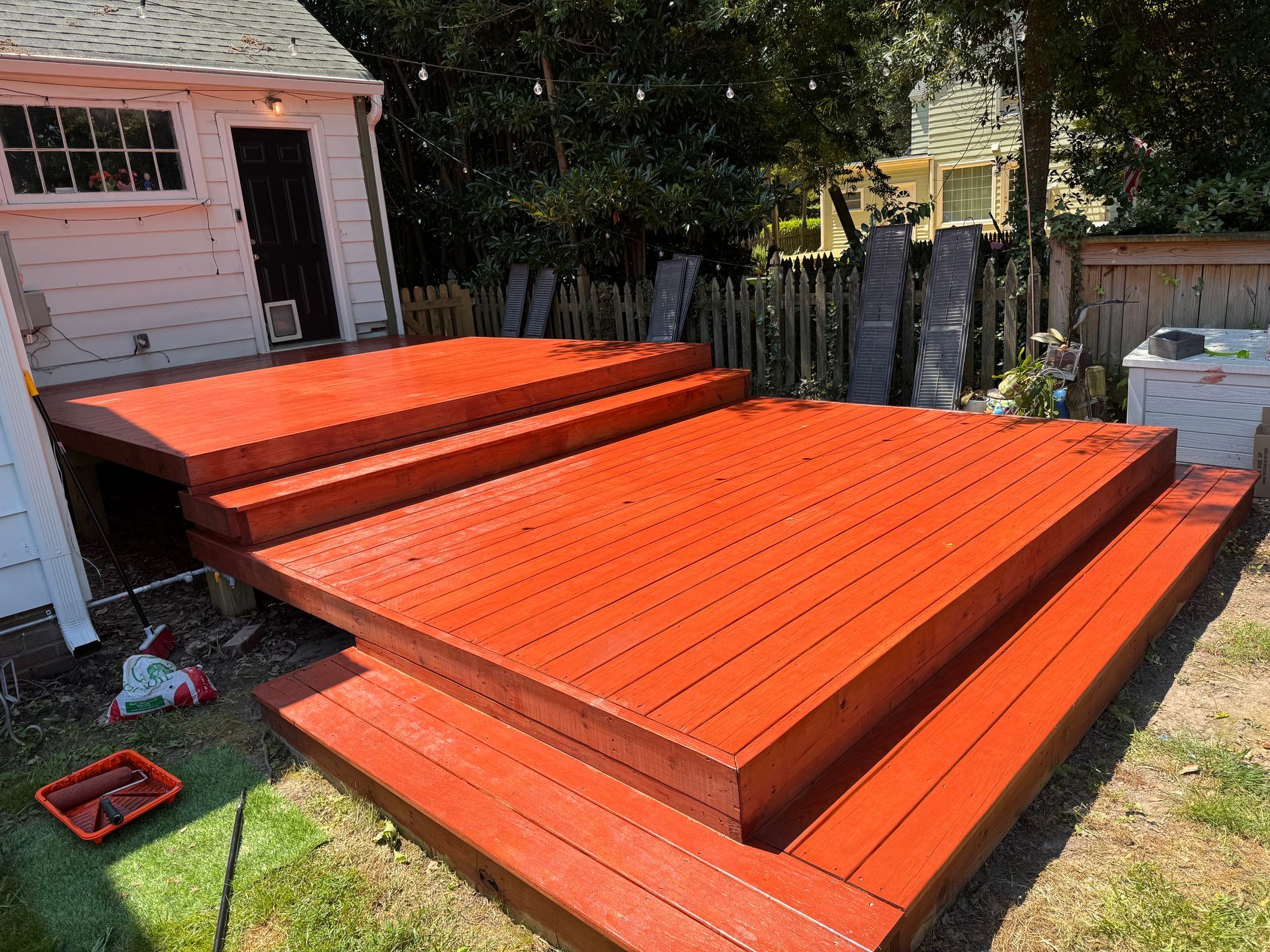 Red-painted wooden deck with steps, in a grassy backyard, adjacent to a white shed.