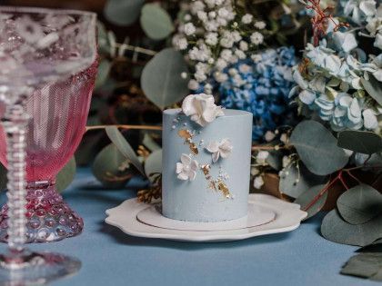 A small, light blue cake with delicate white flowers and gold leaf accents sits on a white plate amid blue florals.