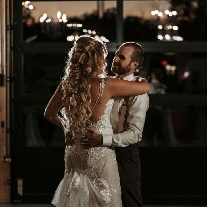 A bride and groom in formal attire share a slow dance together in front of a dark glass door at night.