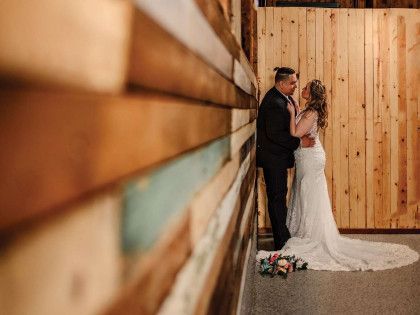 A wedding couple embracing while standing against a rustic wooden wall in an indoor setting.