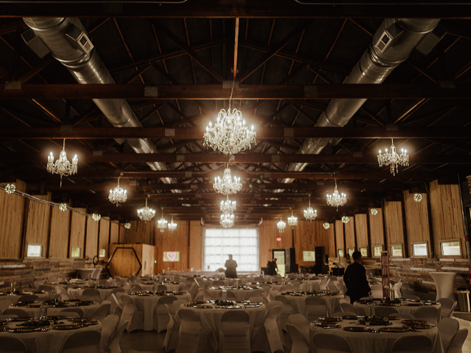 A dimly lit event hall with dark wooden beams, hanging chandeliers, and rows of round tables with white tablecloths.