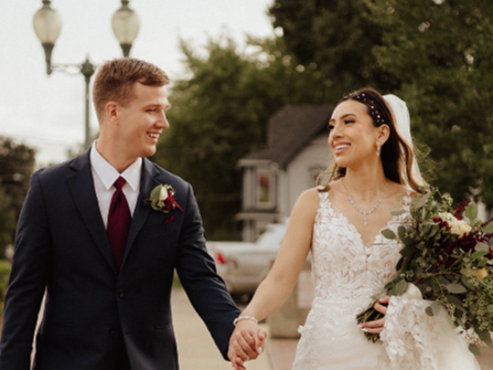A smiling couple in wedding attire walking hand-in-hand outdoors, with the bride holding a floral bouquet.