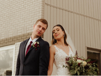 A bride and groom stand back-to-back, eyes closed, against a building wall, holding hands with a floral bouquet.