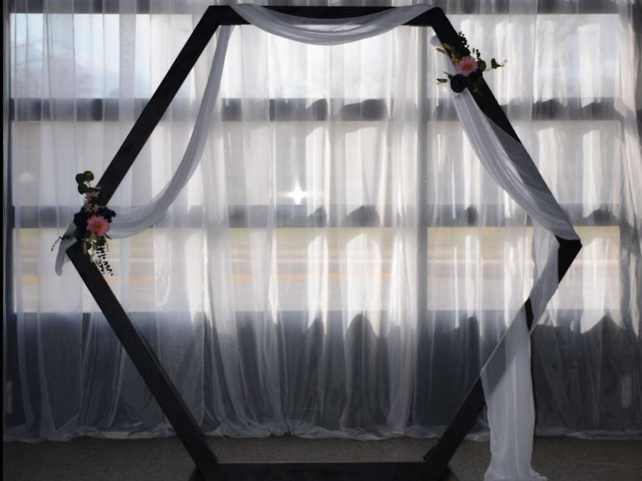 A large, black hexagonal wooden arch decorated with white fabric and small floral accents, set against sheer curtains.