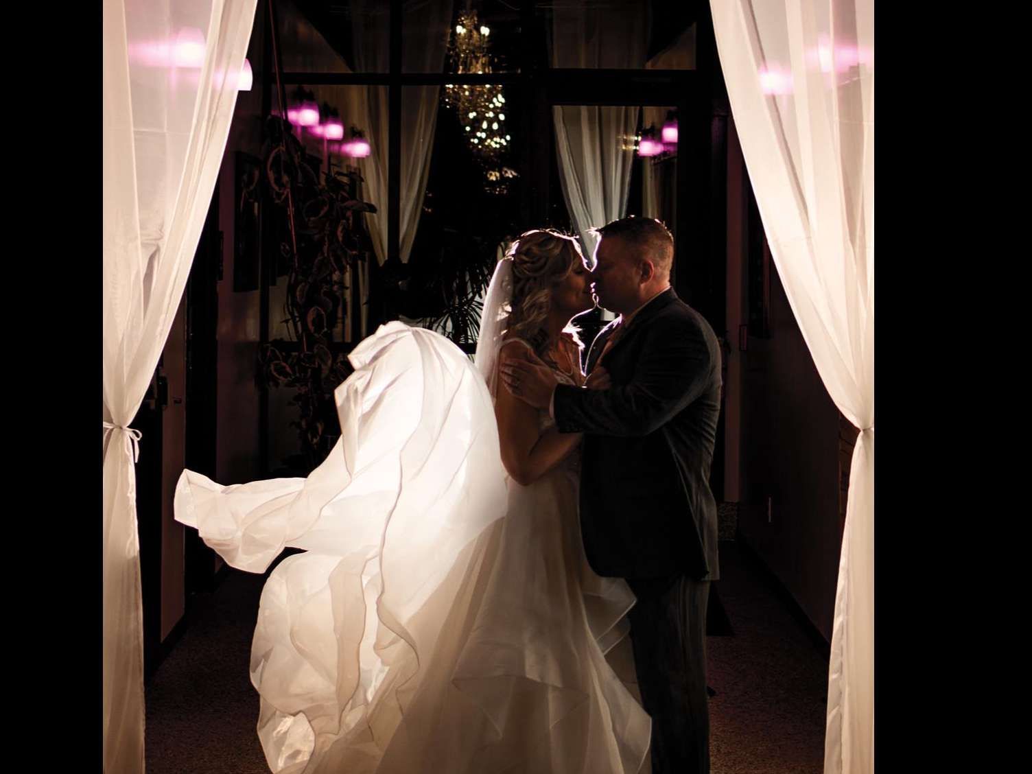 A couple stands in a doorway between white drapes, kissing as the bride’s wedding dress train flares out behind her.