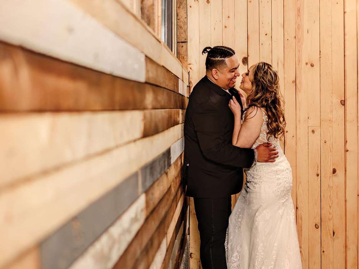 A couple in formal wedding attire smiling at each other in a corner of a rustic room with wood-paneled walls.
