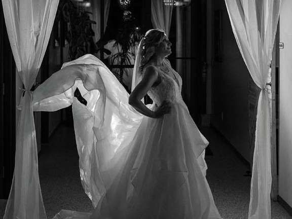 A bride in a white wedding gown stands in a hallway between white drapes, with her veil flowing behind her.