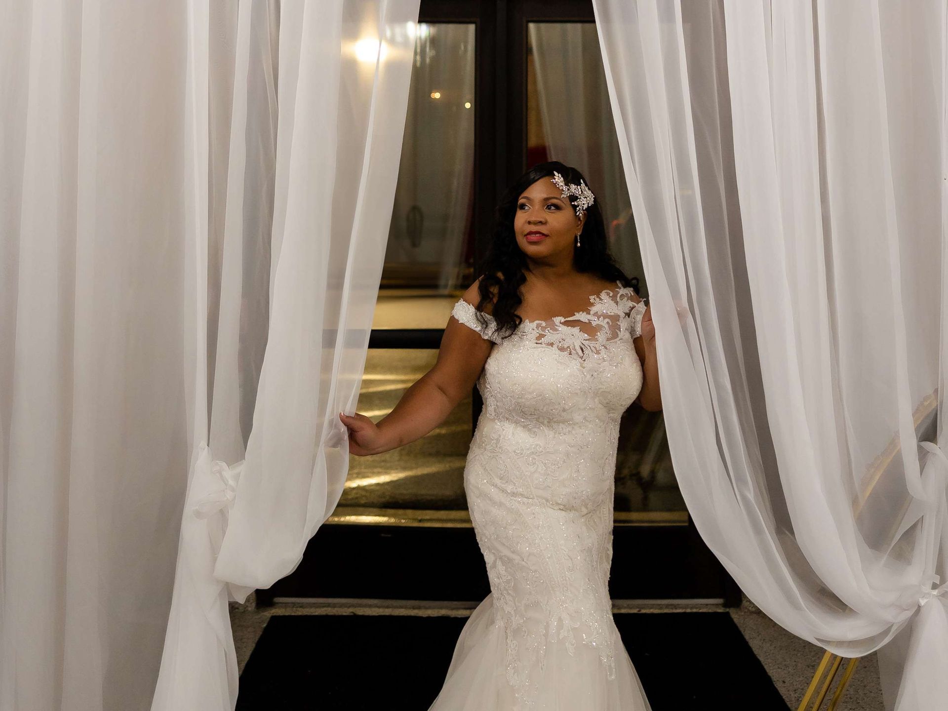 A bride in a white lace mermaid gown stands between flowing white curtains, looking off-camera.