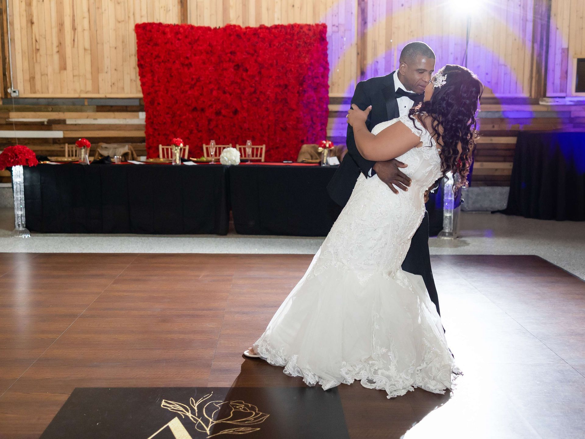 A couple in formal wedding attire kisses on a wooden dance floor in front of a large, vibrant red floral backdrop.