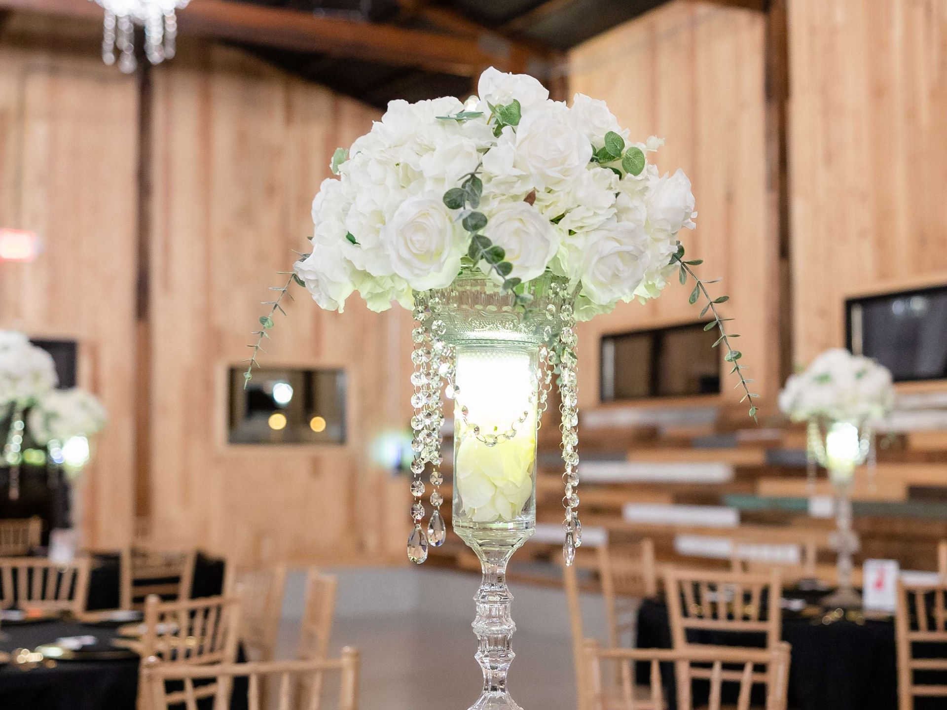 A tall, crystal-style wedding centerpiece with white roses and greenery, set in a rustic wooden event space.