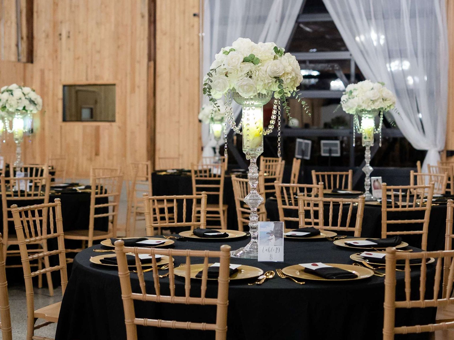 Round tables with black tablecloths, gold chairs, and tall floral centerpieces set up for an indoor event.