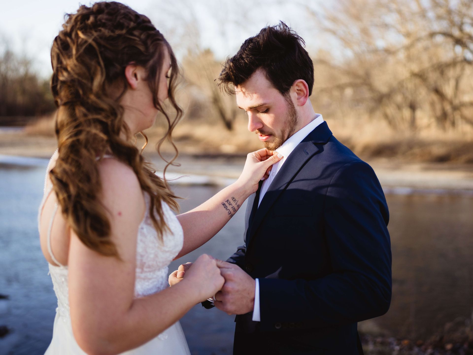A bride in a white wedding dress adjusting a groom's tie outdoors by a river in soft, golden light.