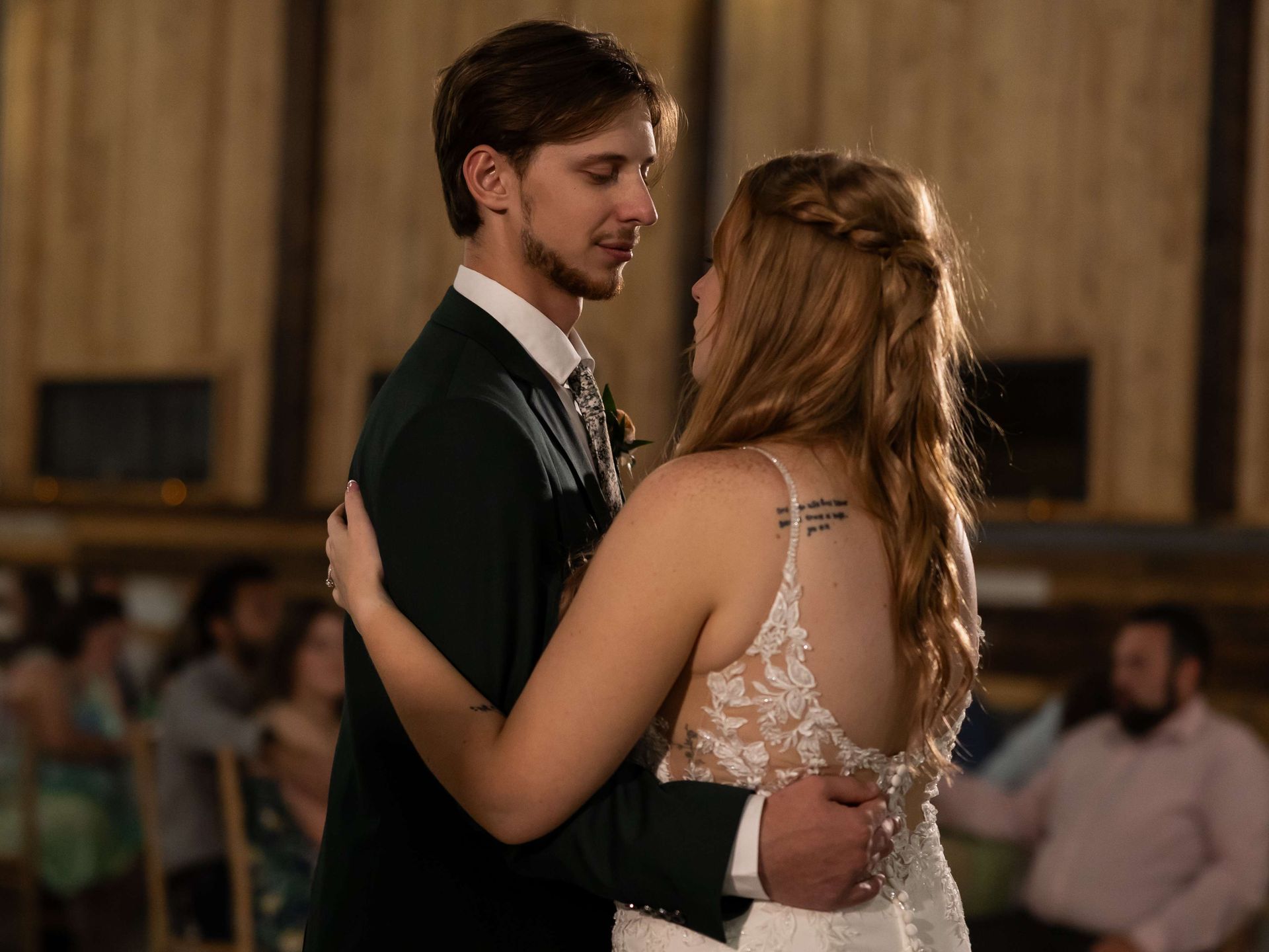 A couple dances at a wedding reception inside a wood-paneled venue, surrounded by guests in the blurred background.