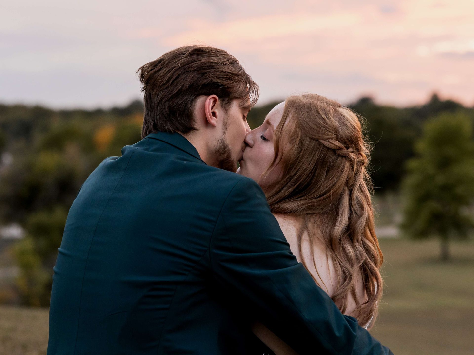 A couple in formal attire embracing and kissing outdoors against a soft, golden-hour sunset background.