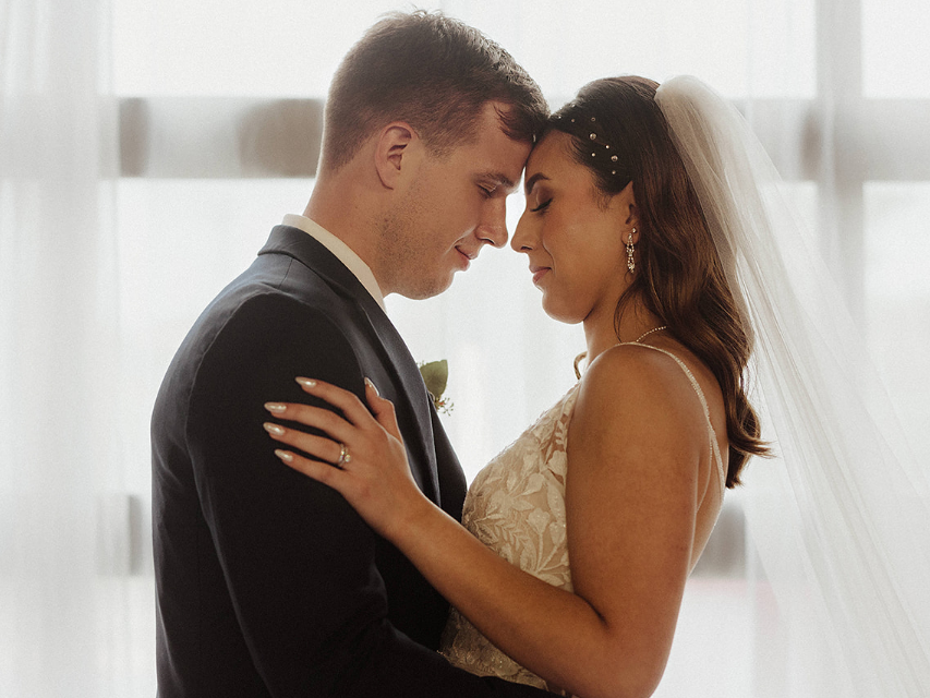 A bride and groom in wedding attire stand face-to-face with eyes closed, foreheads touching, in front of a bright window.