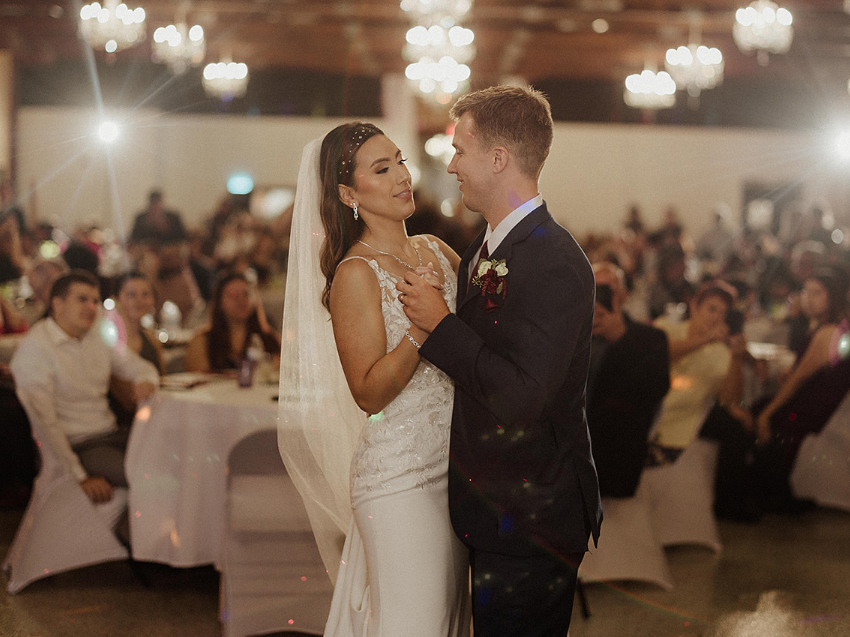 A bride and groom share a wedding dance in a warmly lit ballroom, with guests seated at tables in the blurred background.