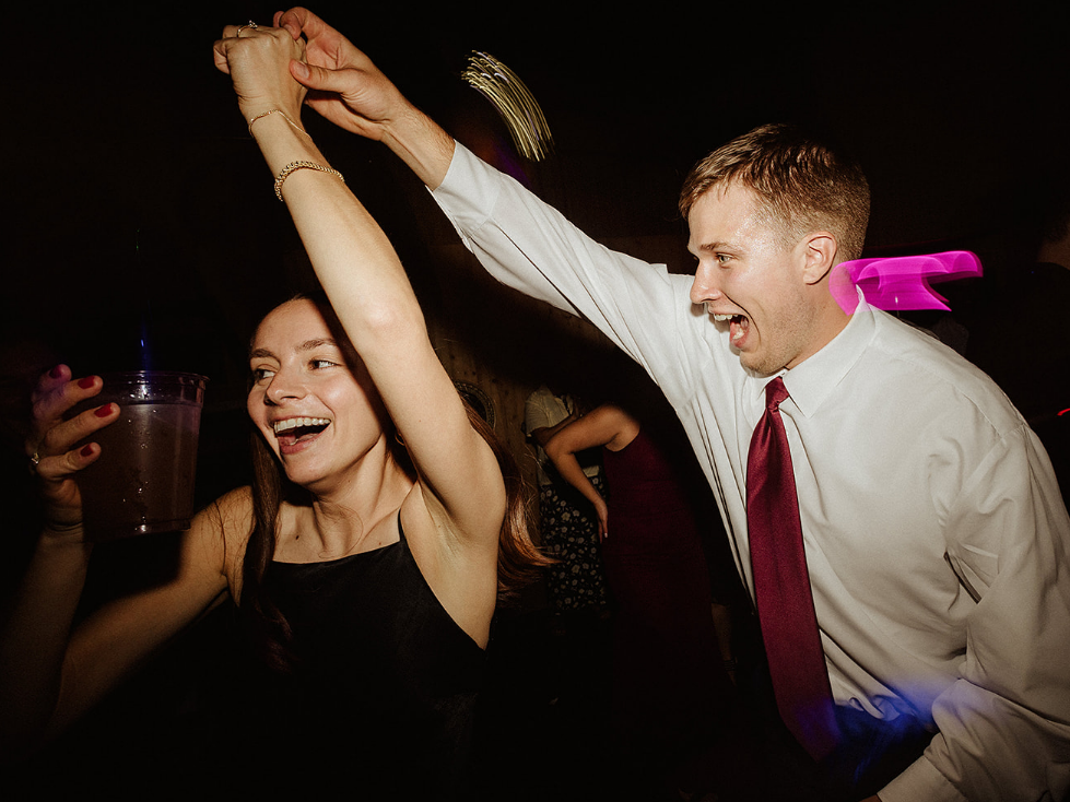 A woman holding a drink and a man in a dress shirt and tie dance together, both laughing in a dimly lit setting.