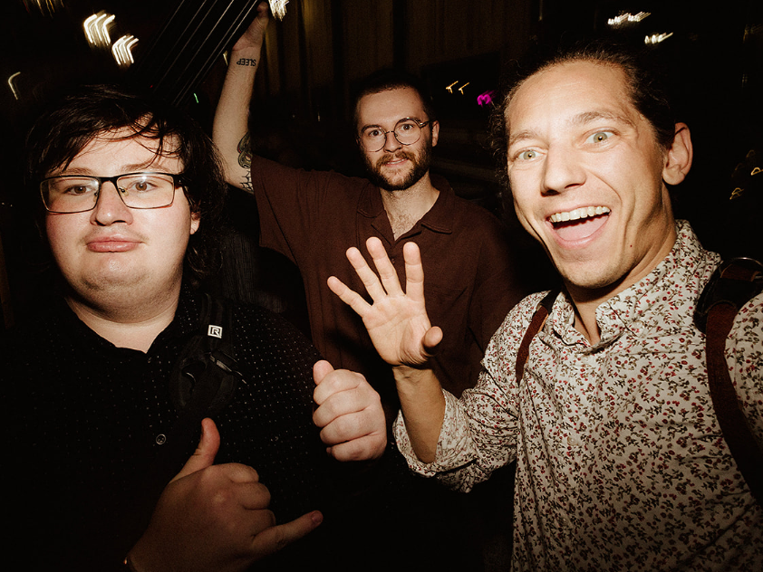 Three people pose for a selfie at night; one smiles with an open hand raised, another gestures with a shaka sign.