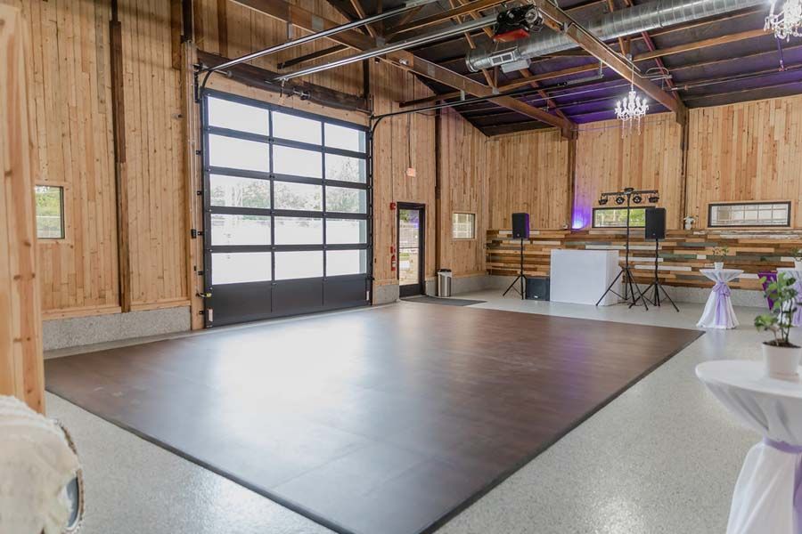 A spacious, rustic event venue with wood-paneled walls, a large glass garage door, and a dark wooden dance floor.