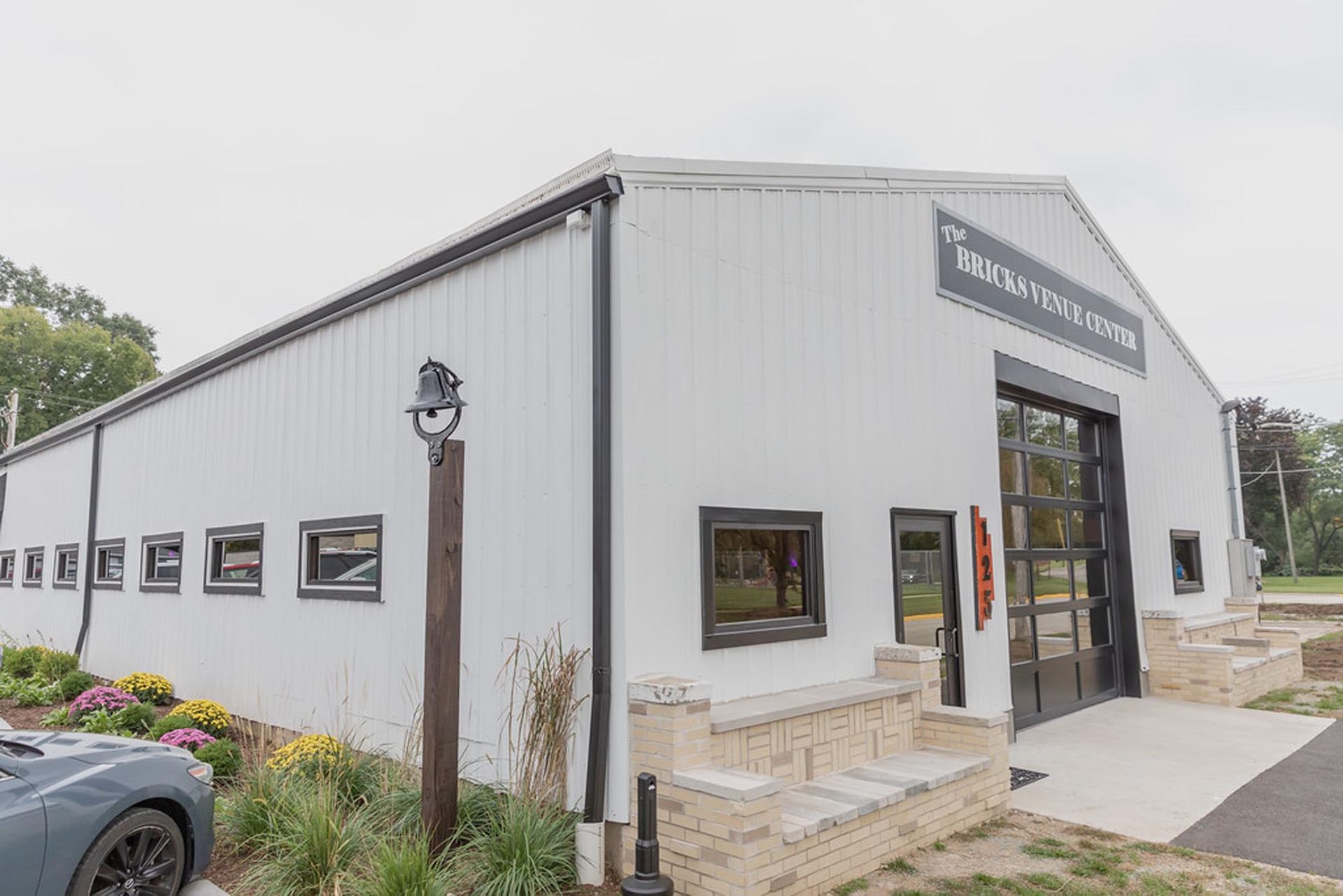 A white metal building with a large glass bay door, signage above, and a stone planter, viewed on an overcast day.