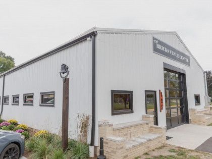 White metal-sided commercial building with a large glass garage door, black trim, and a small flower bed in front.