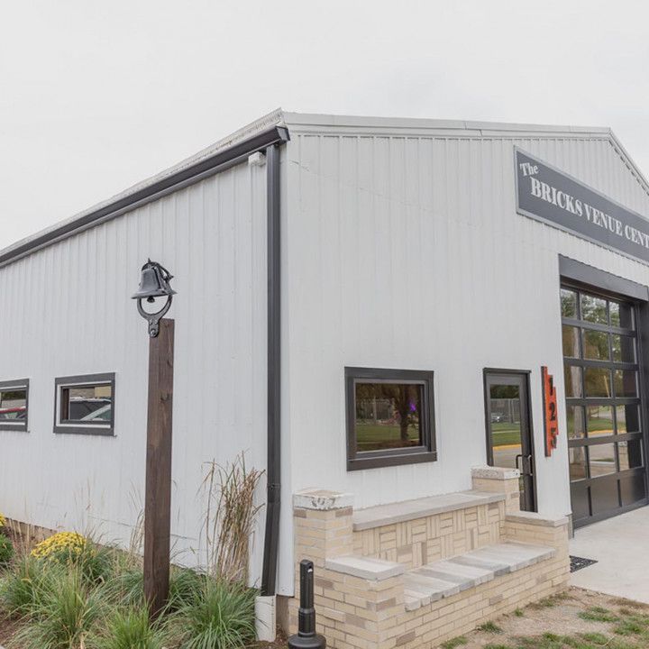 The Bricks Venue building exterior featuring white metal siding, black trim, a large glass bay door, and a brick bench.