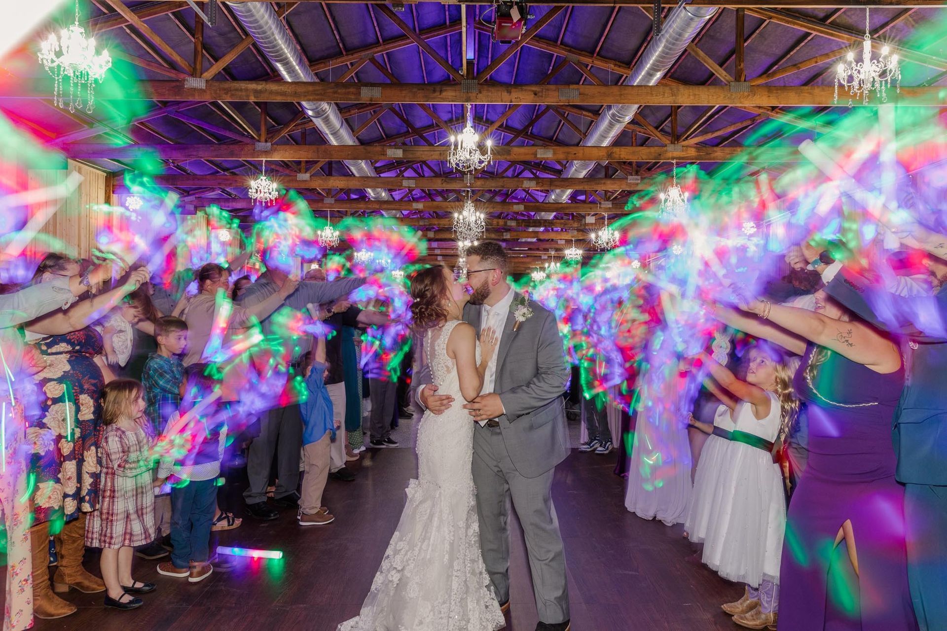 A bride and groom kiss while walking through a cheering crowd holding glowing, multi-colored light sticks in a rustic hall.