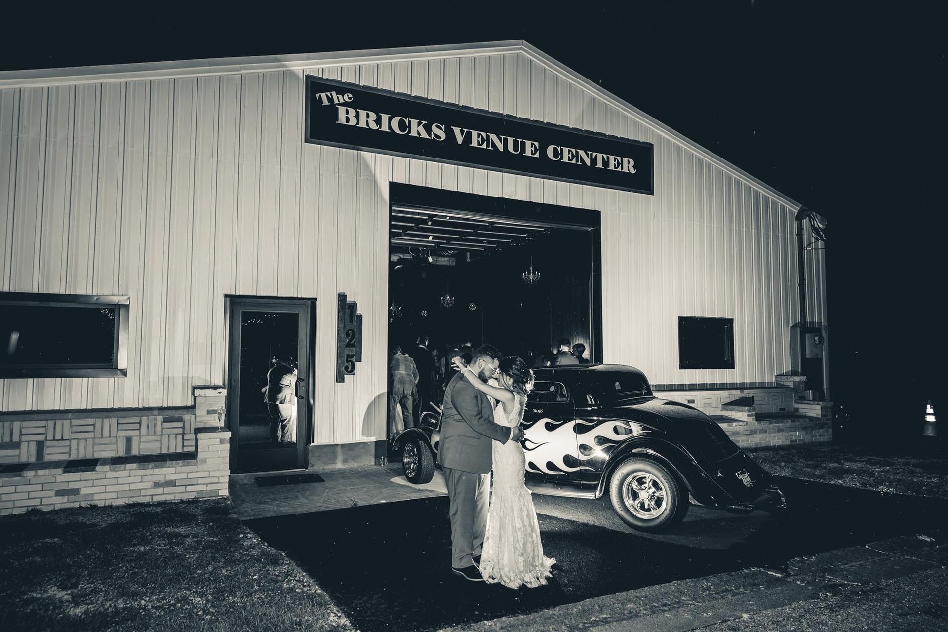 A couple in formal attire embraces in front of a classic car parked outside The Bricks Venue Center at night.