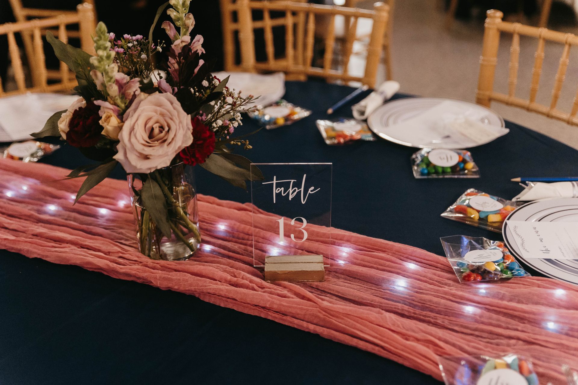 A table at an event featuring a dark blue tablecloth, a pink fabric runner with fairy lights, and a floral centerpiece.