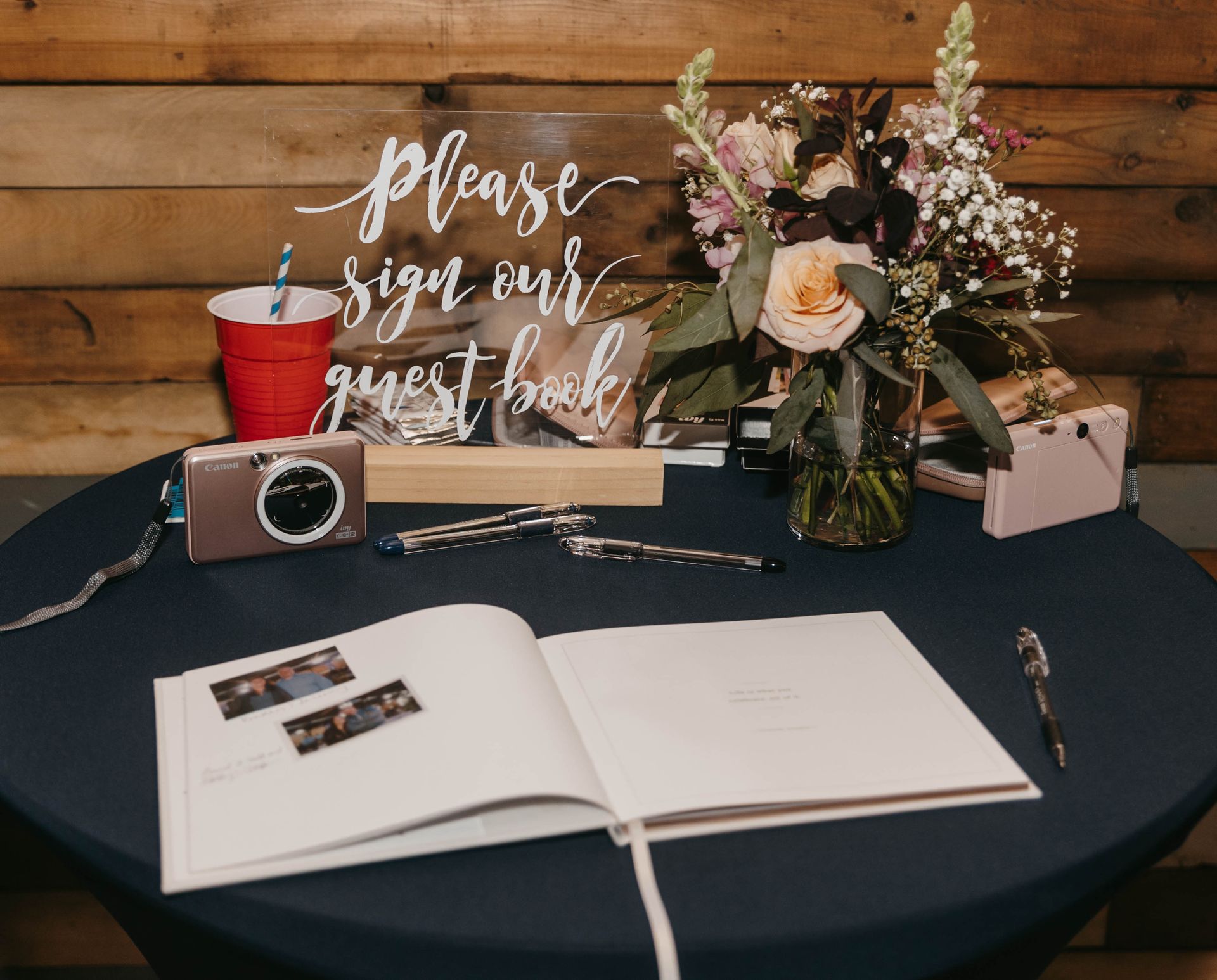 A guest book table with an acrylic sign, floral arrangement, instant camera, and photos on a dark blue tablecloth.