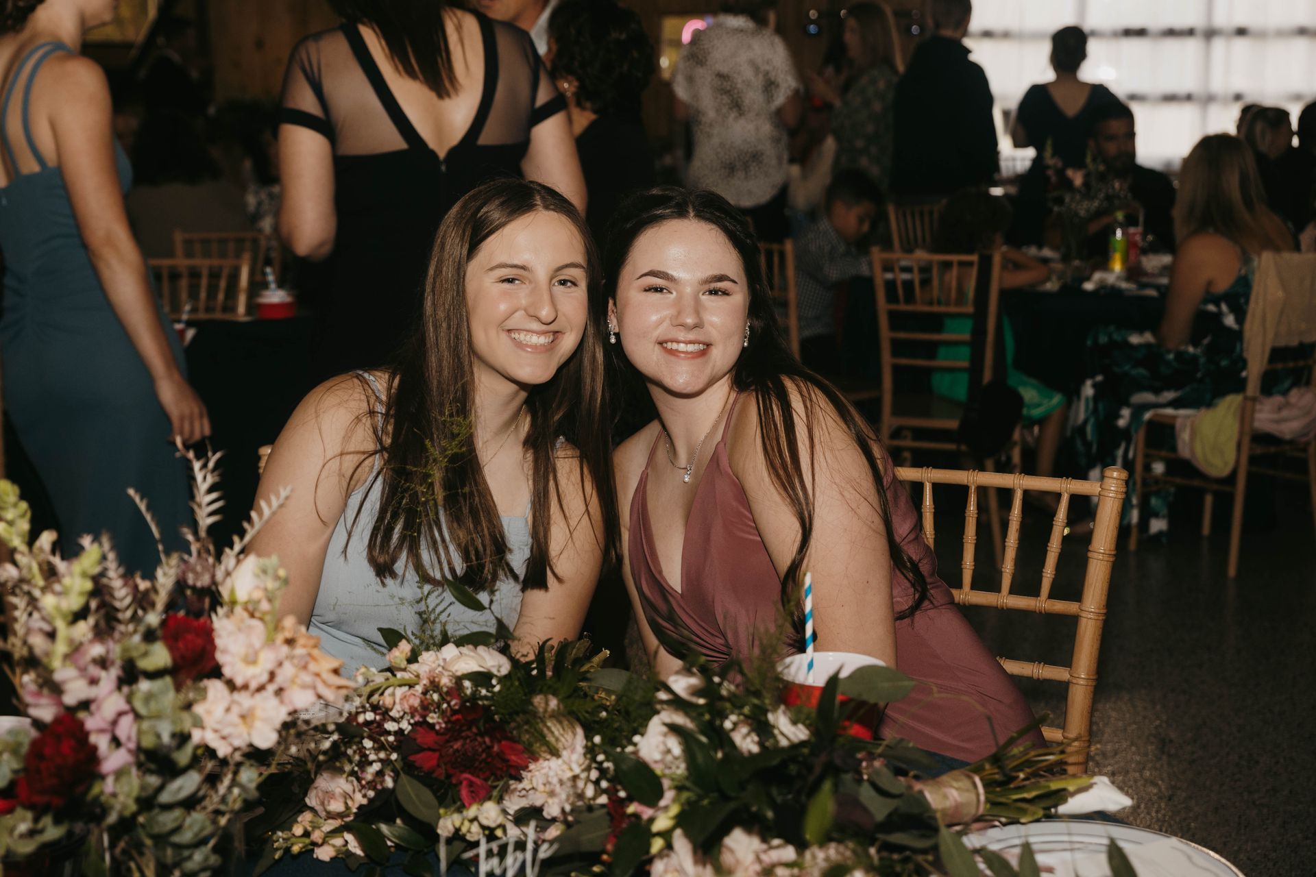 Two smiling friends sit at a table decorated with flowers in a dimly lit banquet hall, facing the camera together.