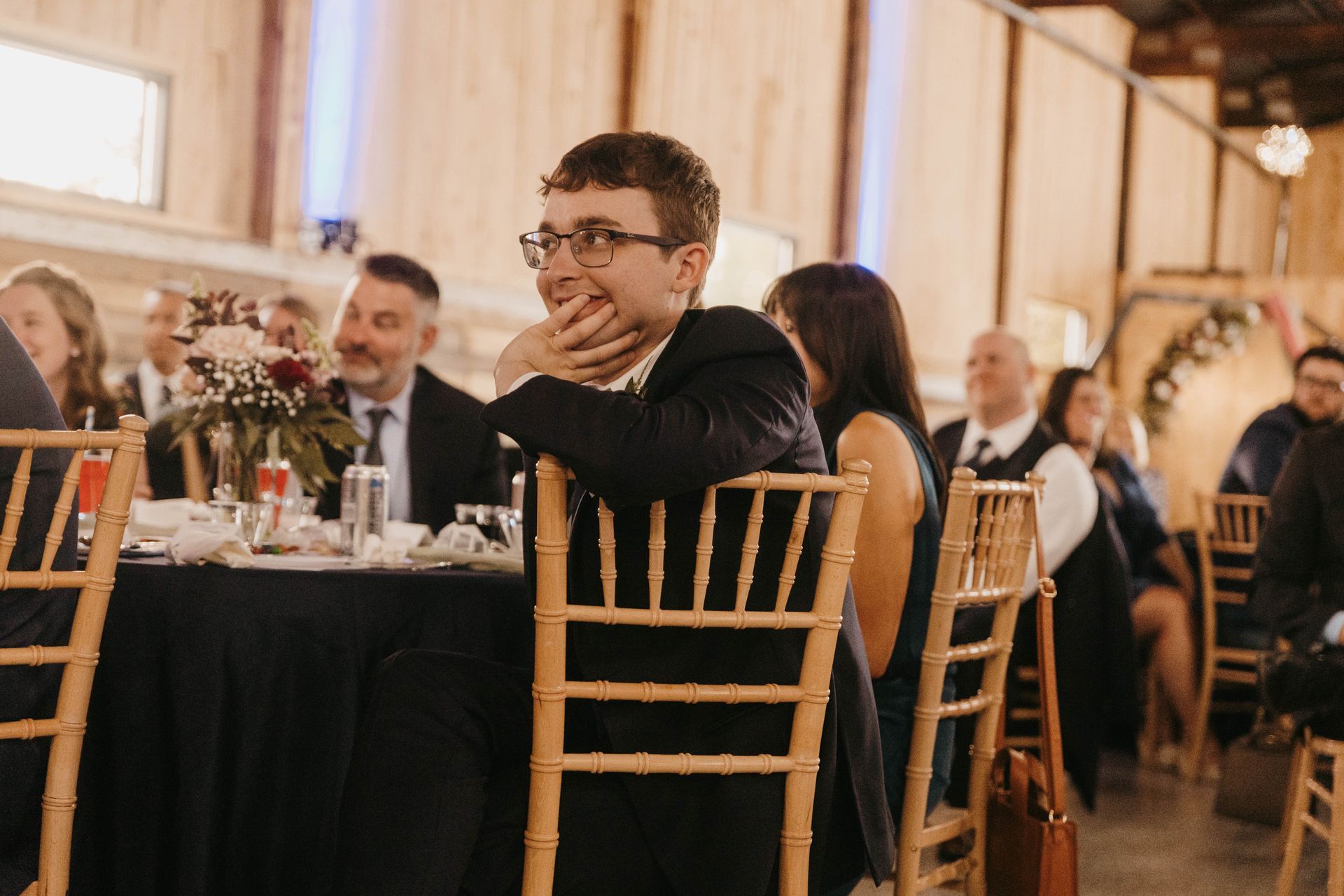 A person in a suit sits at a table during an event, smiling with their hand on their chin while others eat in the background.