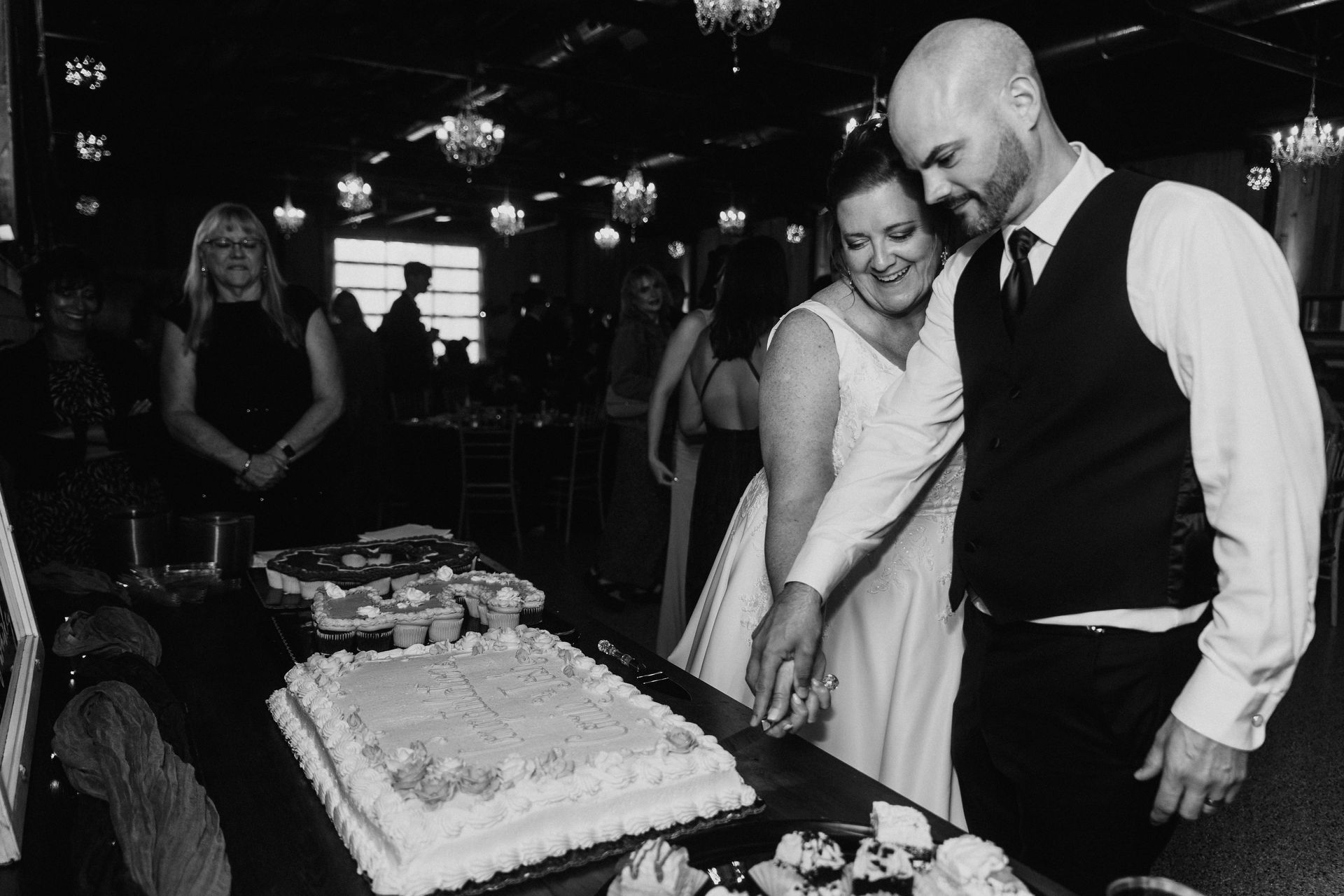A couple cuts a large, rectangular wedding cake together at a dim, festive reception.
