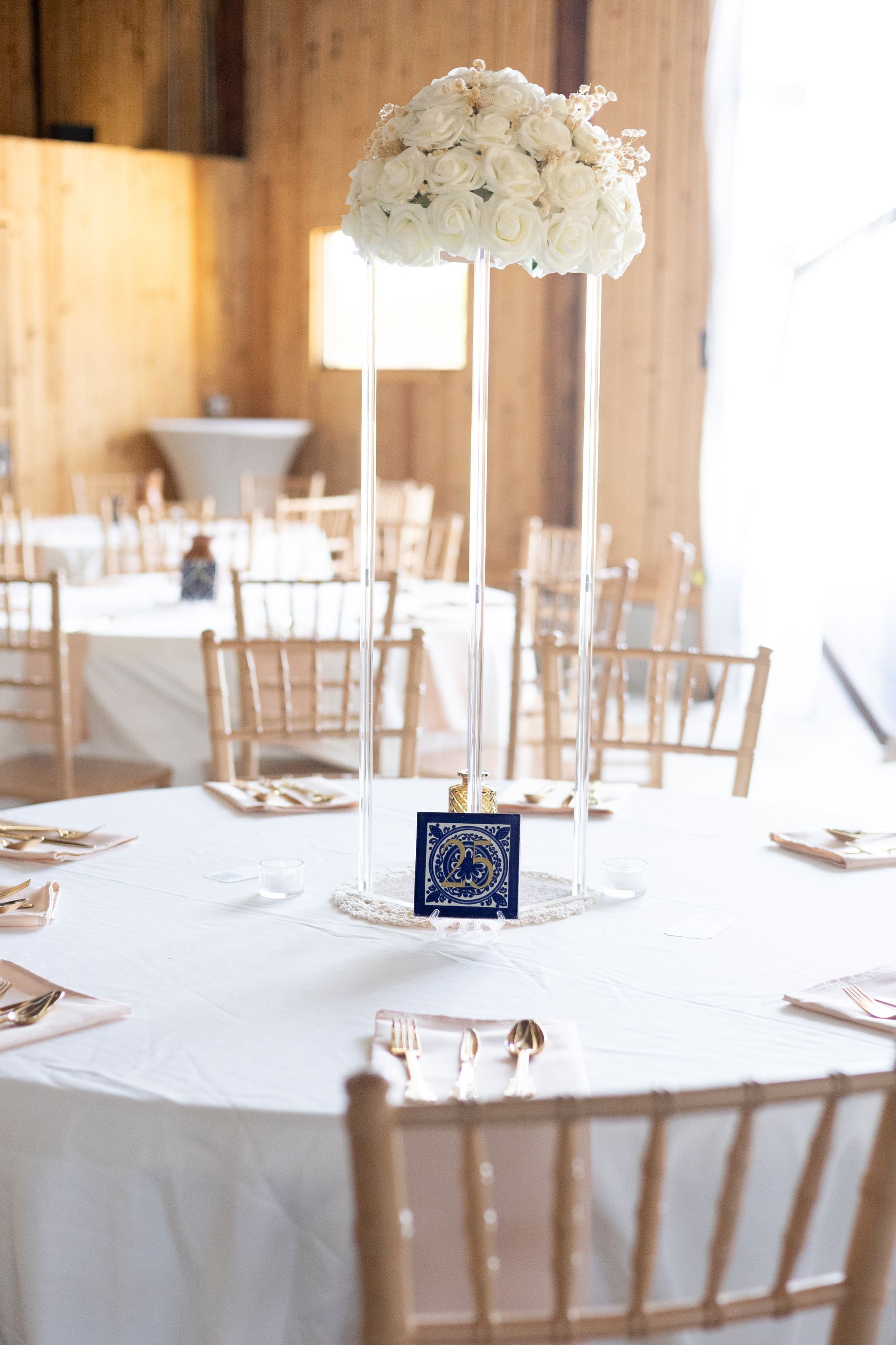 A tall, clear acrylic centerpiece holding a large bouquet of white flowers on a white-clothed table in a wooden venue.