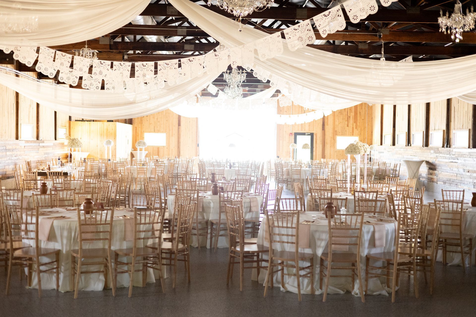 A large rustic event venue filled with round tables, gold chairs, white tablecloths, and ceiling drapes with chandeliers.