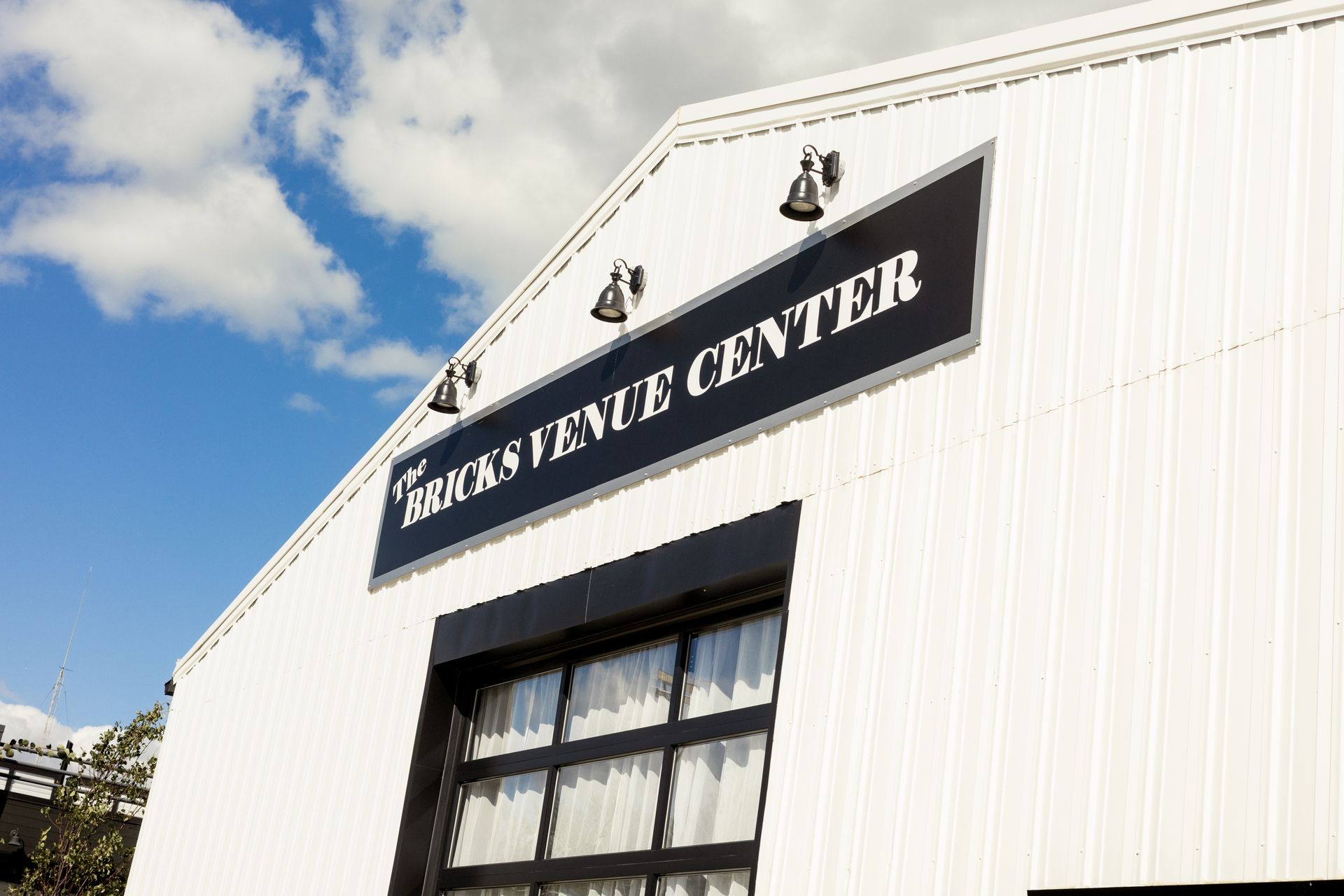 Exterior of The Bricks Venue Center, a white building with a black sign and a grid-style glass door under a blue sky.