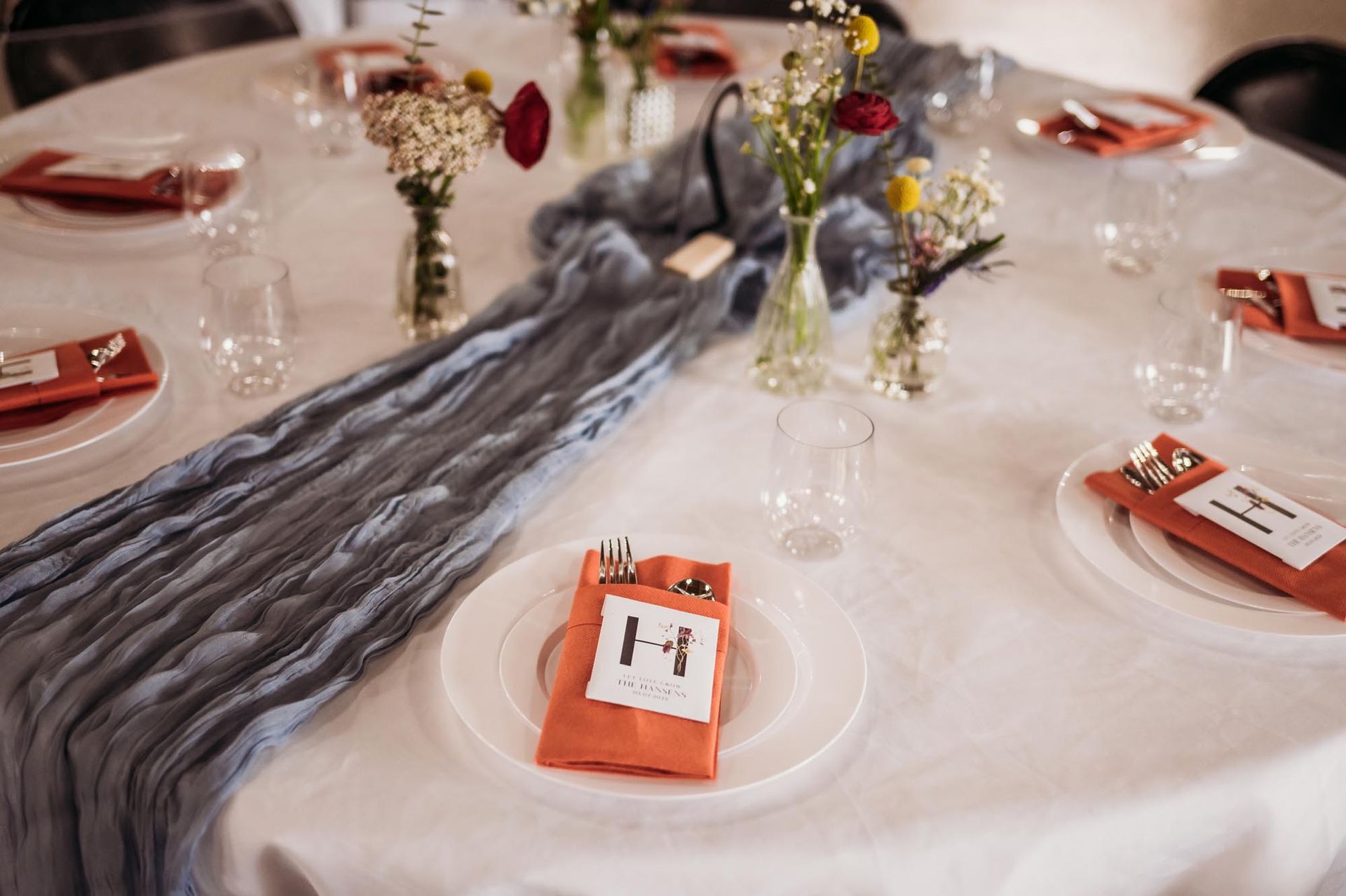 Round wedding table with a blue runner, terracotta napkins, white plates, and small floral arrangements.