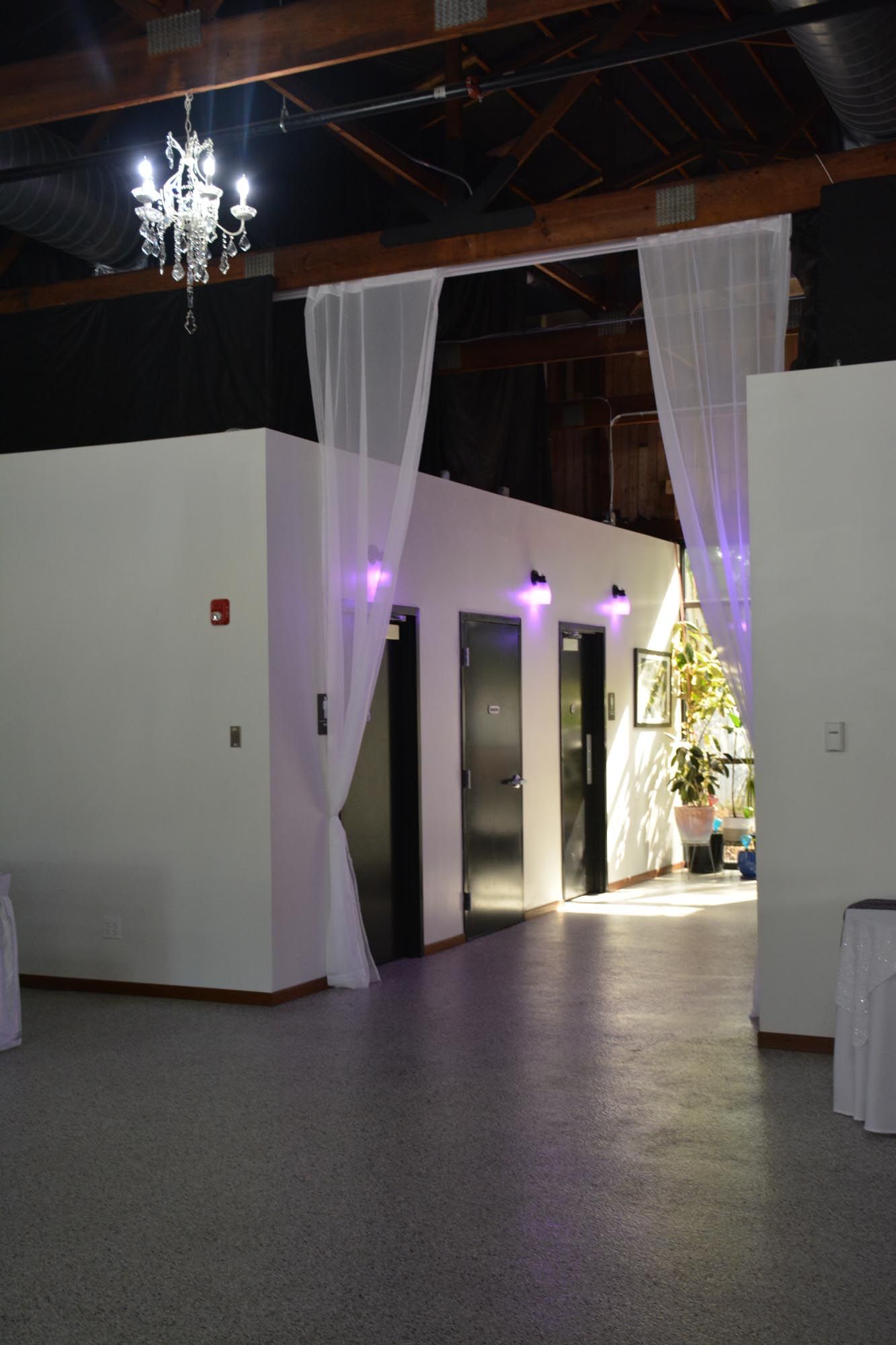 A bright event space with white walls, a chandelier, and a walkway draped with white curtains leading to dark doorways.