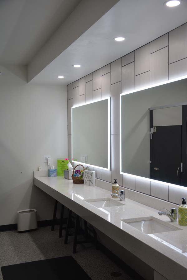 A modern bathroom vanity with two illuminated rectangular mirrors, a white countertop, two sinks, and tiled walls.