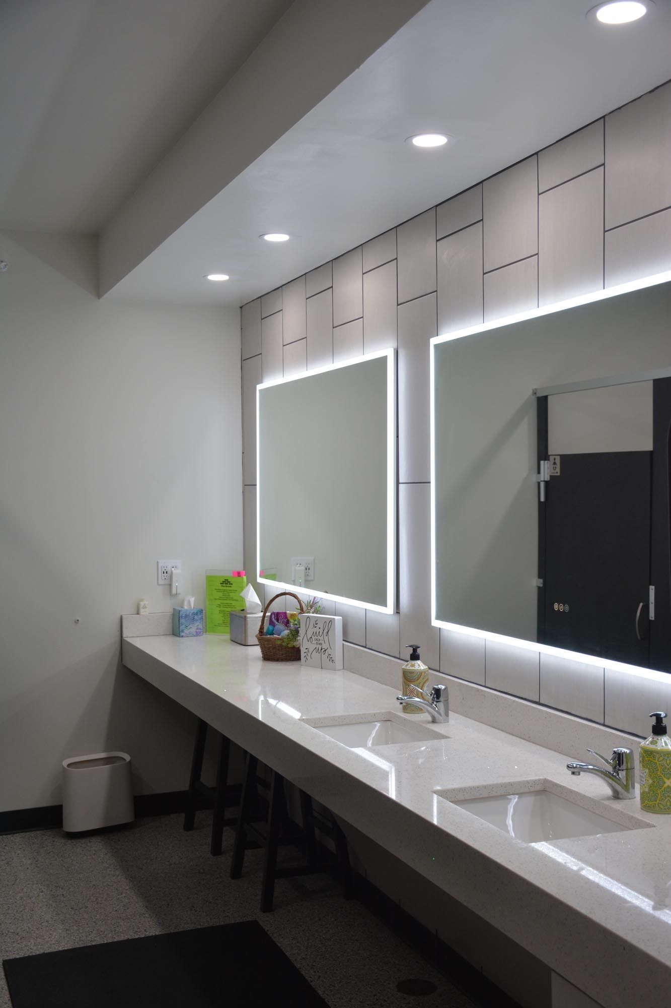 Modern public restroom with two backlit mirrors over a long, white quartz vanity with twin sinks and under-counter stools.