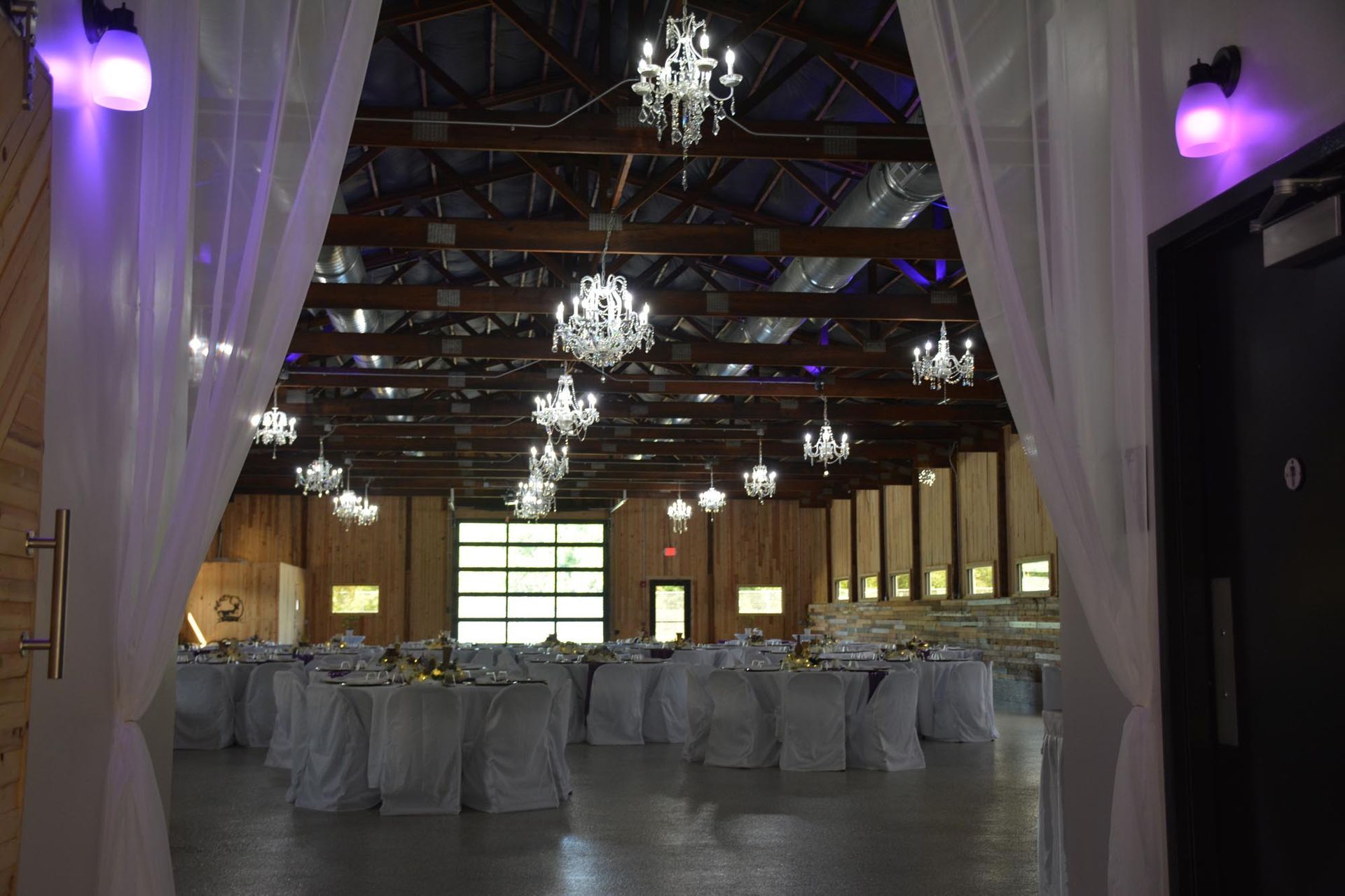 An elegant indoor banquet hall with crystal chandeliers, round tables with white linens, and warm, dark wood rafters.
